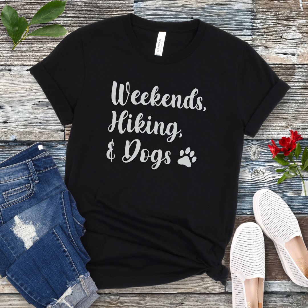 A black t-shirt with the text 'Weekends, Hiking, & Dogs' text in playful, feminine script and a paw print, displayed on a wooden surface alongside denim jeans and white sneakers.