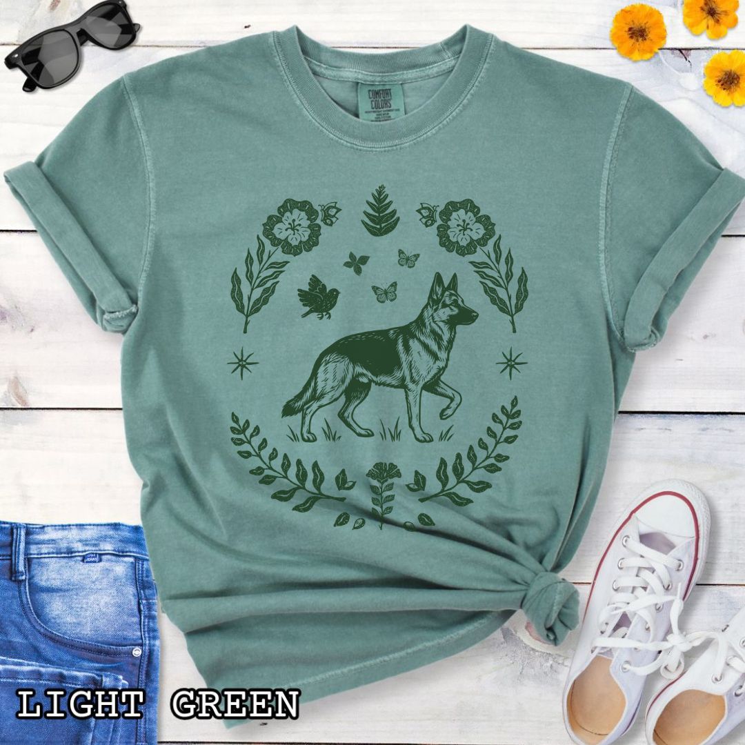 Green Comfort Colors t-shirt for German Shepherd lovers with monochrome linocut-style design of a German Shepherd dog surrounded by floral botanical elements, shown on a wooden surface with jeans and sneakers.