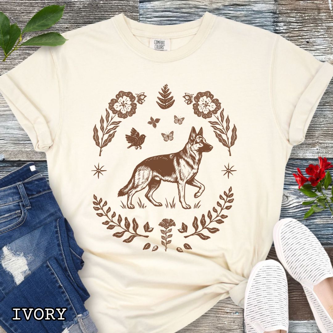 Beige Comfort Colors t-shirt for German Shepherd lovers with monochrome linocut-style design of a German Shepherd dog surrounded by floral botanical elements, shown on a wooden surface with jeans and sneakers.