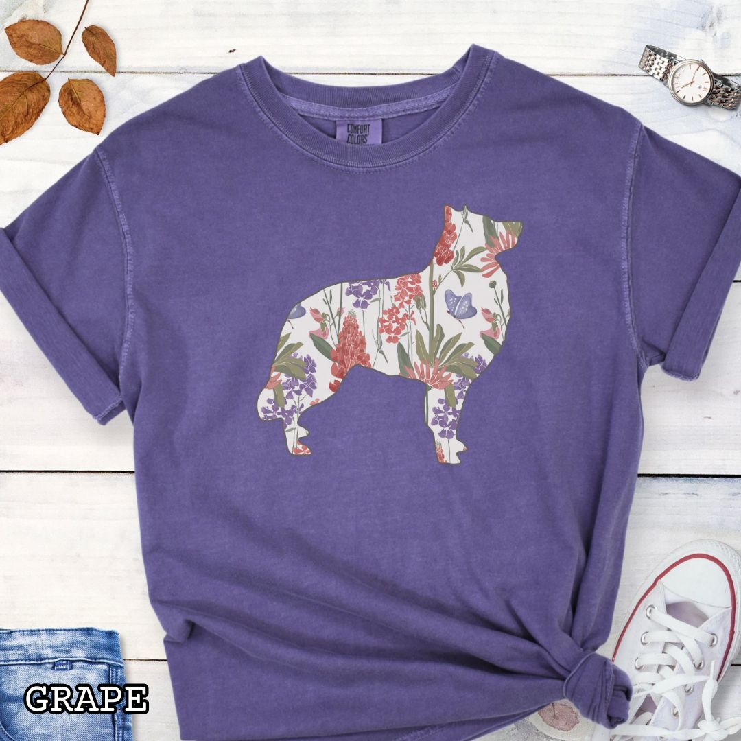 Purple Comfort Colors t-shirt with a floral-filled silhouette of a Belgian Sheepdog/Tervuren, accompanied by a pair of rolled-up blue jeans and a pair of white shoes on a wooden surface.