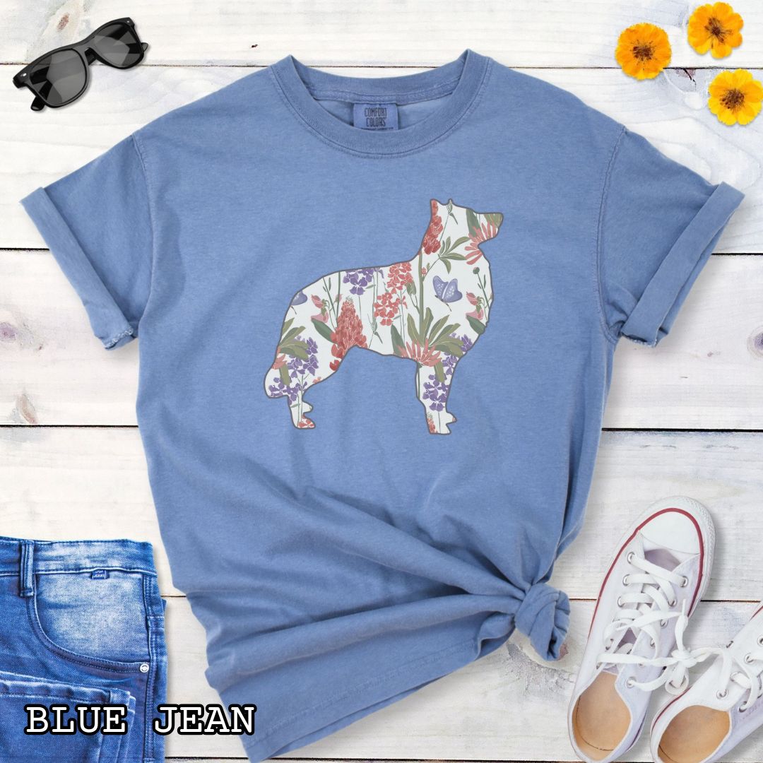 Blue Comfort Colors t-shirt with a floral-filled silhouette of a Belgian Sheepdog/Tervuren, accompanied by a pair of rolled-up blue jeans and a pair of white shoes on a wooden surface.
