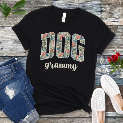 A black Bella Canvas 3001 unisex short sleeve T-shirt with floral-filled text 'DOG grammy' printed on the front, displayed on a wooden surface alongside denim shorts and white sneakers.