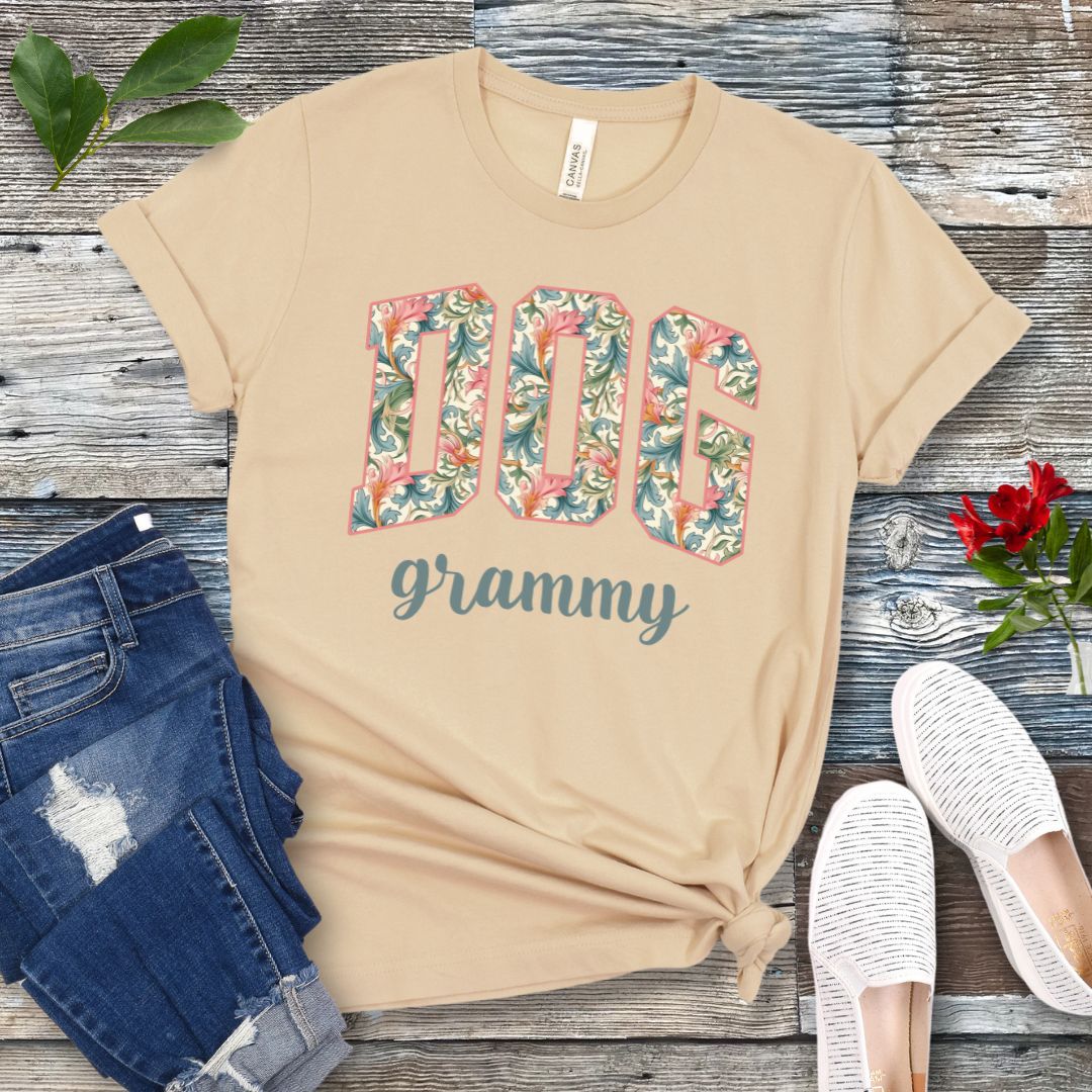 A cream Bella Canvas 3001 unisex short sleeve T-shirt with floral-filled text 'DOG grammy' printed on the front, displayed on a wooden surface alongside denim shorts and white sneakers.