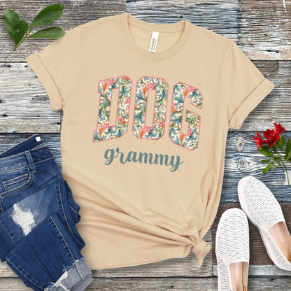 A cream Bella Canvas 3001 unisex short sleeve T-shirt with floral-filled text 'DOG grammy' printed on the front, displayed on a wooden surface alongside denim shorts and white sneakers.