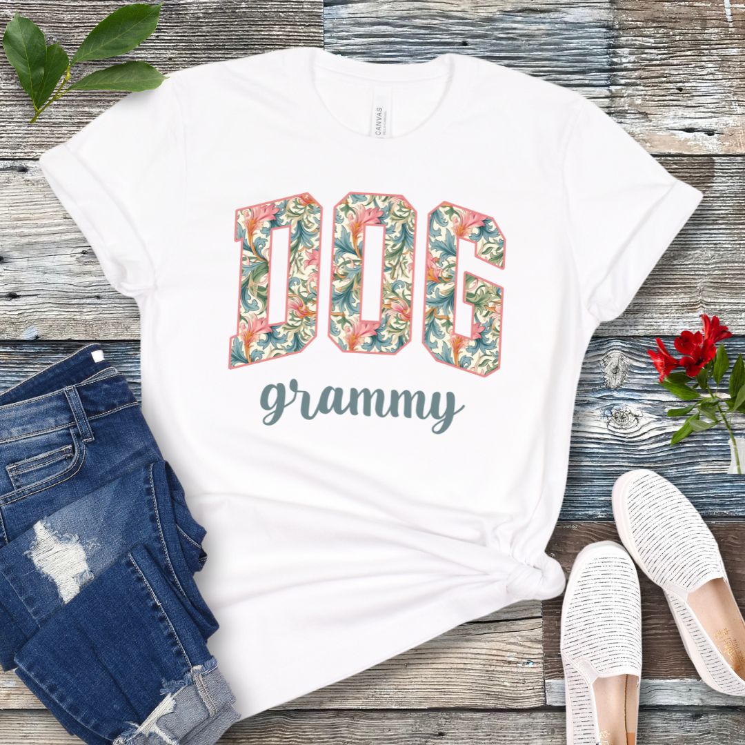 A white Bella Canvas 3001 unisex short sleeve T-shirt with floral-filled text 'DOG grammy' printed on the front, displayed on a wooden surface alongside denim shorts and white sneakers.