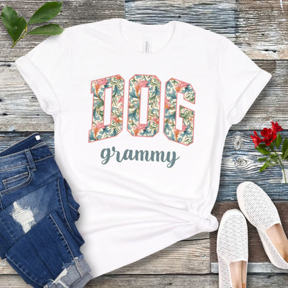 A white Bella Canvas 3001 unisex short sleeve T-shirt with floral-filled text 'DOG grammy' printed on the front, displayed on a wooden surface alongside denim shorts and white sneakers.
