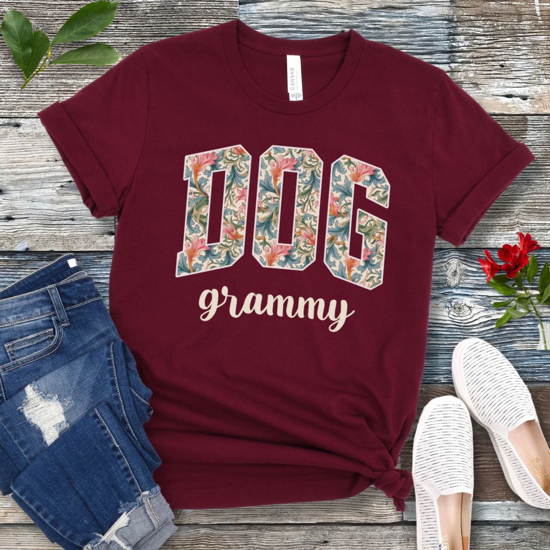 A maroon Bella Canvas 3001 unisex short sleeve T-shirt with floral-filled text 'DOG grammy' printed on the front, displayed on a wooden surface alongside denim shorts and white sneakers.
