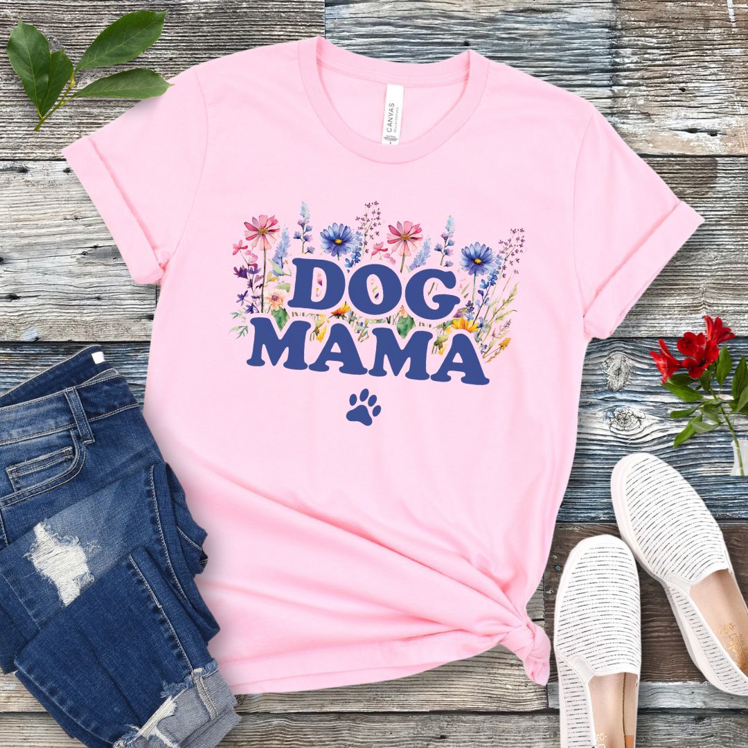 A pink t-shirt with a vibrant wildflower design surrounding the text 'DOG MAMA', and a paw print underneath. The shirt is laid out flat with a pair of blue jeans and white shoes in the background.