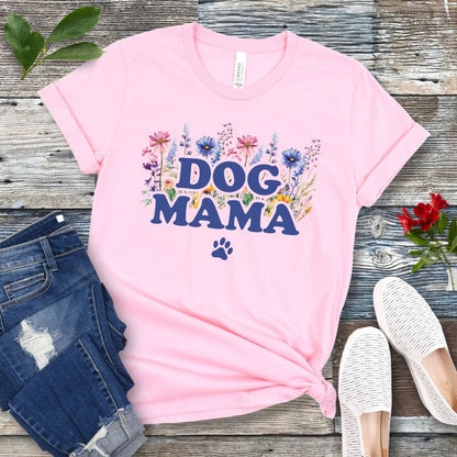 A pink t-shirt with a vibrant wildflower design surrounding the text 'DOG MAMA', and a paw print underneath. The shirt is laid out flat with a pair of blue jeans and white shoes in the background.