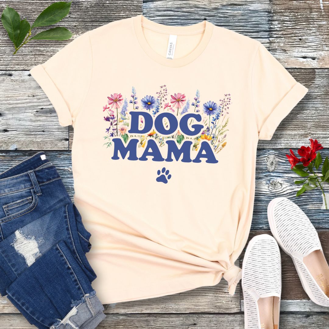 A beige t-shirt with a vibrant wildflower design surrounding the text 'DOG MAMA', and a paw print underneath. The shirt is laid out flat with a pair of blue jeans and white shoes in the background.