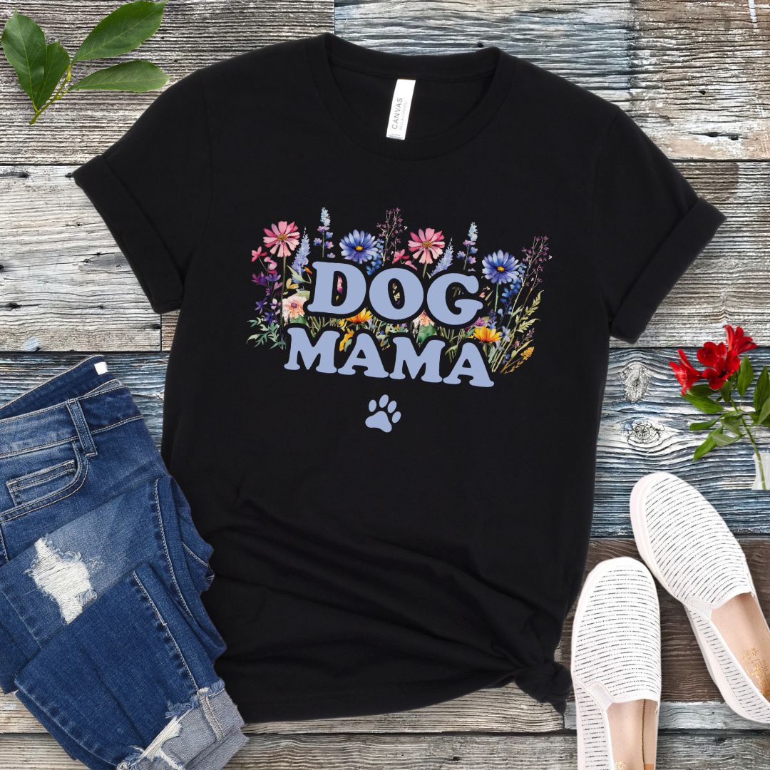 A black t-shirt with a vibrant wildflower design surrounding the text 'DOG MAMA', and a paw print underneath. The shirt is laid out flat with a pair of blue jeans and white shoes in the background.