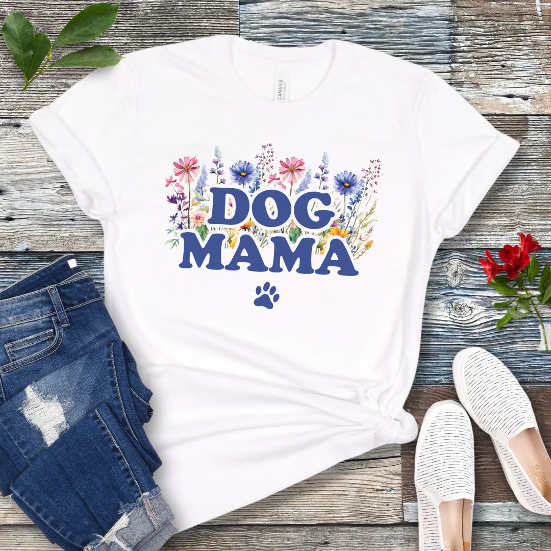 A white t-shirt with a vibrant wildflower design surrounding the text 'DOG MAMA', and a paw print underneath. The shirt is laid out flat with a pair of blue jeans and white shoes in the background.