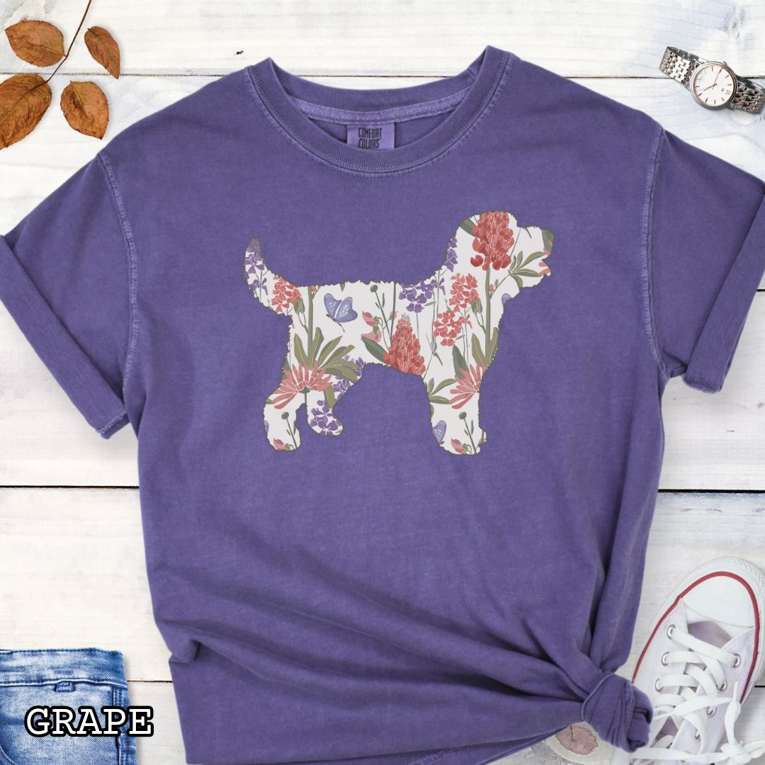 Purple Comfort Colors t-shirt with a floral-filled silhouette of a Doodle dog, accompanied by a pair of rolled-up blue jeans and a pair of white shoes on a wooden surface.