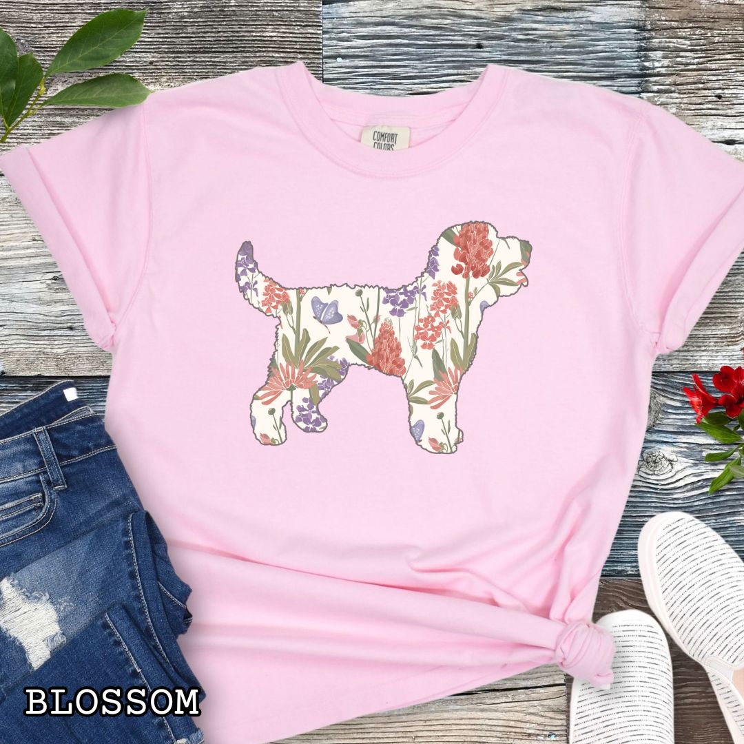 Pink Comfort Colors t-shirt with a floral-filled silhouette of a Doodle dog, accompanied by a pair of rolled-up blue jeans and a pair of white shoes on a wooden surface.