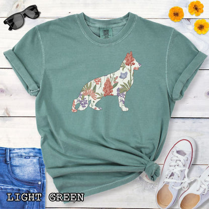 Green Comfort Colors t-shirt with a floral-filled silhouette of a German Shepherd, accompanied by a pair of rolled-up blue jeans and a pair of white shoes on a wooden surface.