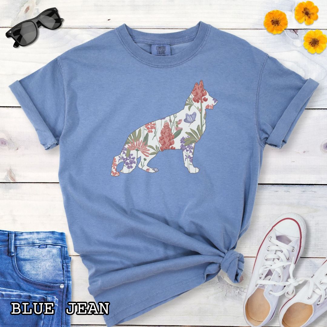 Blue Comfort Colors t-shirt with a floral-filled silhouette of a German Shepherd, accompanied by a pair of rolled-up blue jeans and a pair of white shoes on a wooden surface.