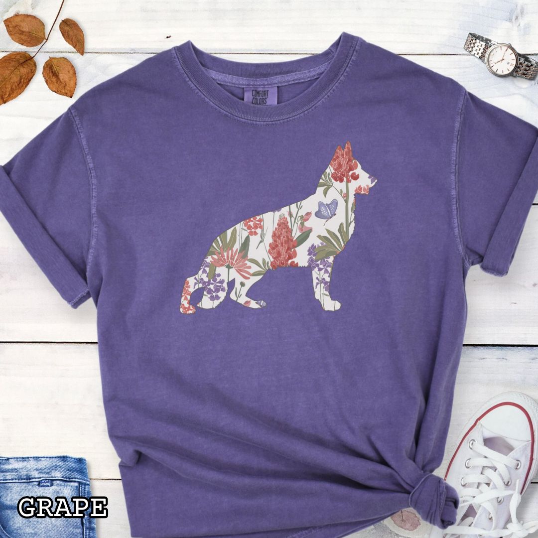 Purple Comfort Colors t-shirt with a floral-filled silhouette of a German Shepherd, accompanied by a pair of rolled-up blue jeans and a pair of white shoes on a wooden surface.