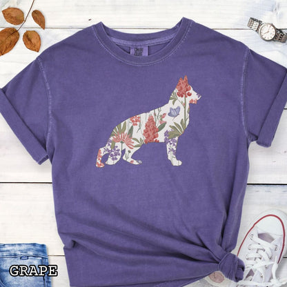 Purple Comfort Colors t-shirt with a floral-filled silhouette of a German Shepherd, accompanied by a pair of rolled-up blue jeans and a pair of white shoes on a wooden surface.
