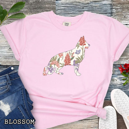 Pink (Blossom) Comfort Colors t-shirt with a floral-filled silhouette of a German Shepherd, accompanied by a pair of rolled-up blue jeans and a pair of white shoes on a wooden surface.