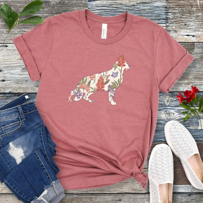 A mauve t-shirt with a floral-filled silhouette of a German Shepherd, accompanied by a pair of rolled-up blue jeans and a pair of white shoes on a wooden surface.