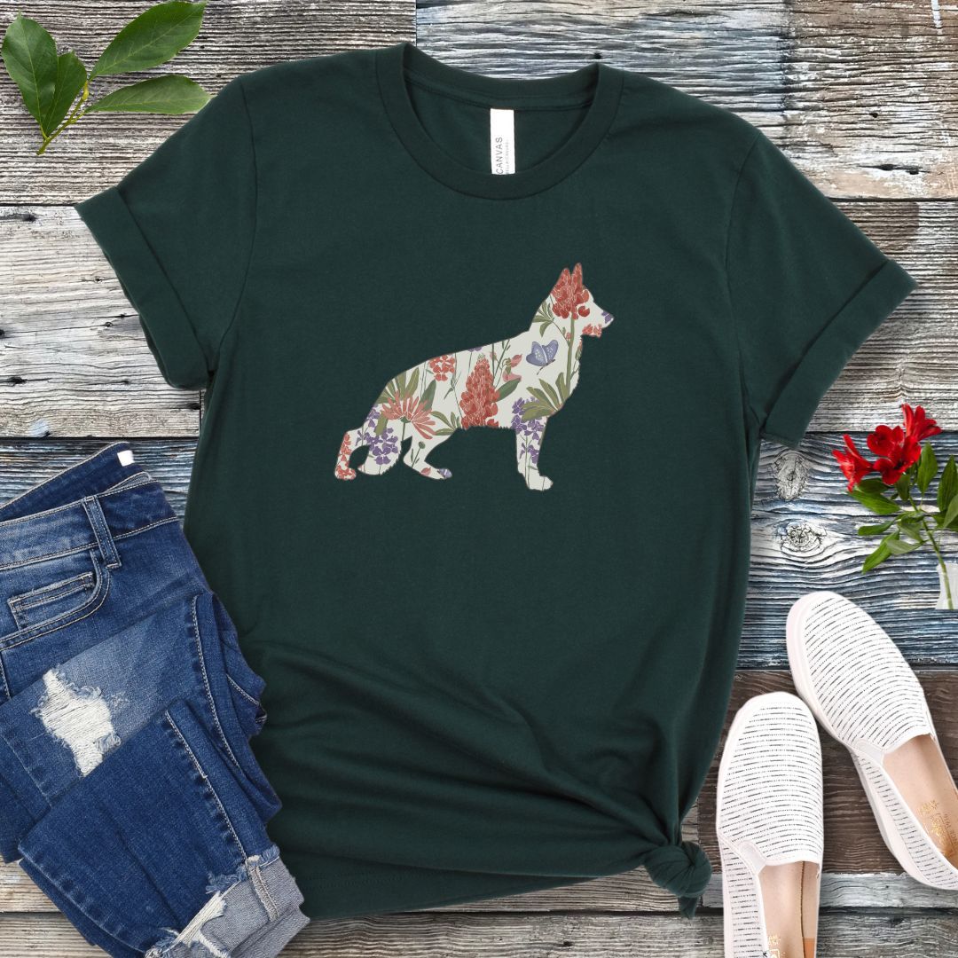 A forest green t-shirt with a floral-filled silhouette of a German Shepherd, accompanied by a pair of rolled-up blue jeans and a pair of white shoes on a wooden surface.