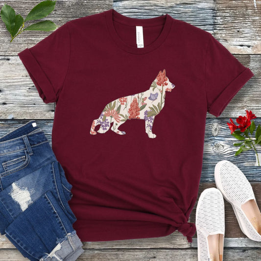 A maroon t-shirt with a floral-filled silhouette of a German Shepherd, accompanied by a pair of rolled-up blue jeans and a pair of white shoes on a wooden surface.