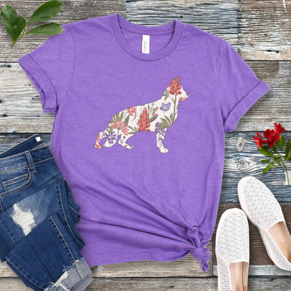 A purple t-shirt with a floral-filled silhouette of a German Shepherd, accompanied by a pair of rolled-up blue jeans and a pair of white shoes on a wooden surface.