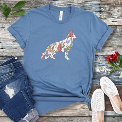 A steel blue t-shirt with a floral-filled silhouette of a German Shepherd, accompanied by a pair of rolled-up blue jeans and a pair of white shoes on a wooden surface.