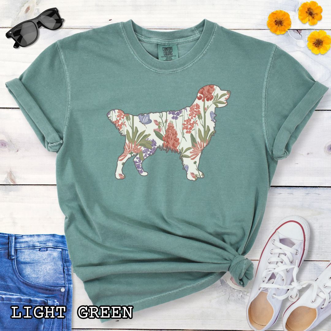 Light green Comfort Colors t-shirt with a floral-filled silhouette of a Golden Retriever, accompanied by a pair of rolled-up blue jeans and a pair of white shoes on a wooden surface.