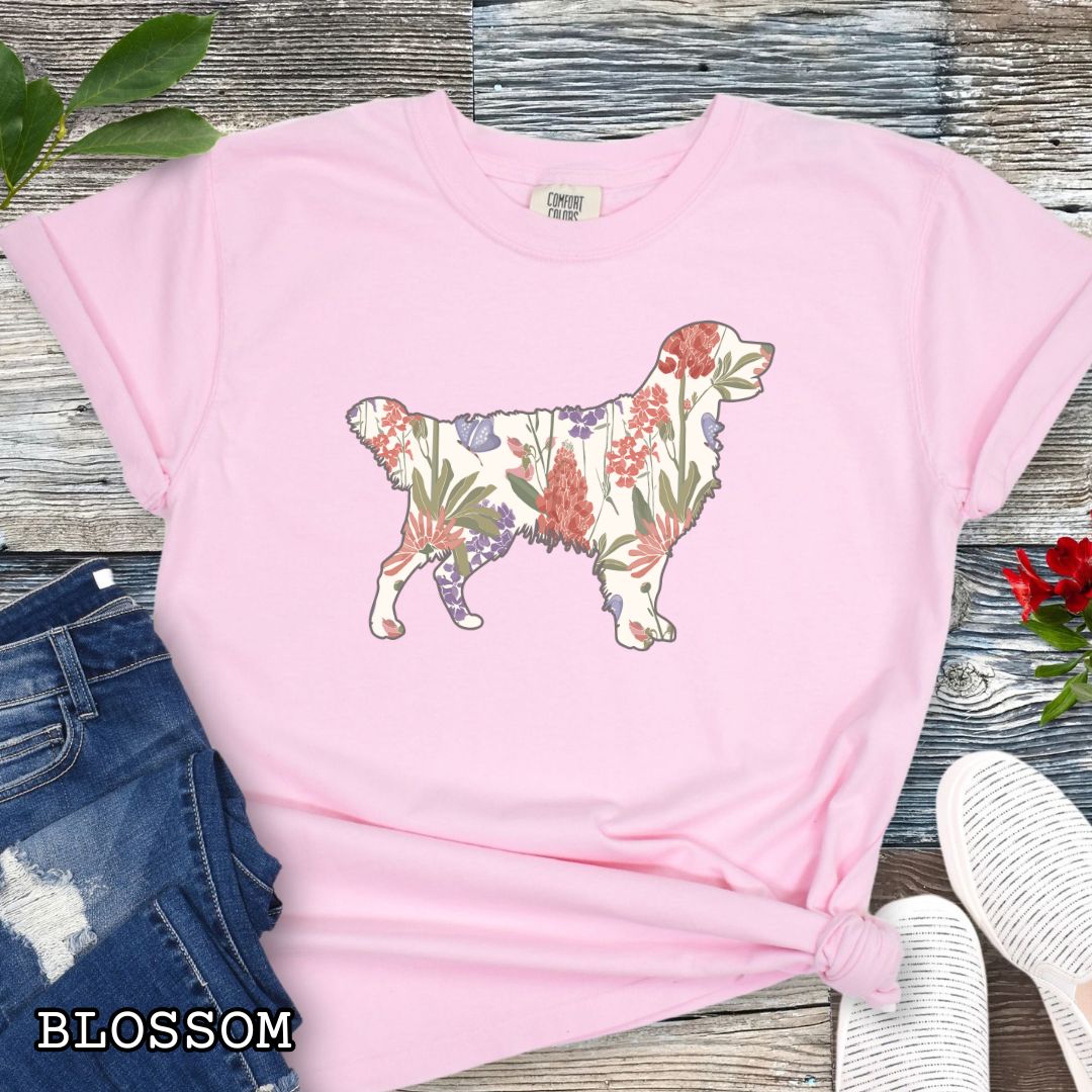 Pink Comfort Colors t-shirt with a floral-filled silhouette of a Golden Retriever, accompanied by a pair of rolled-up blue jeans and a pair of white shoes on a wooden surface.