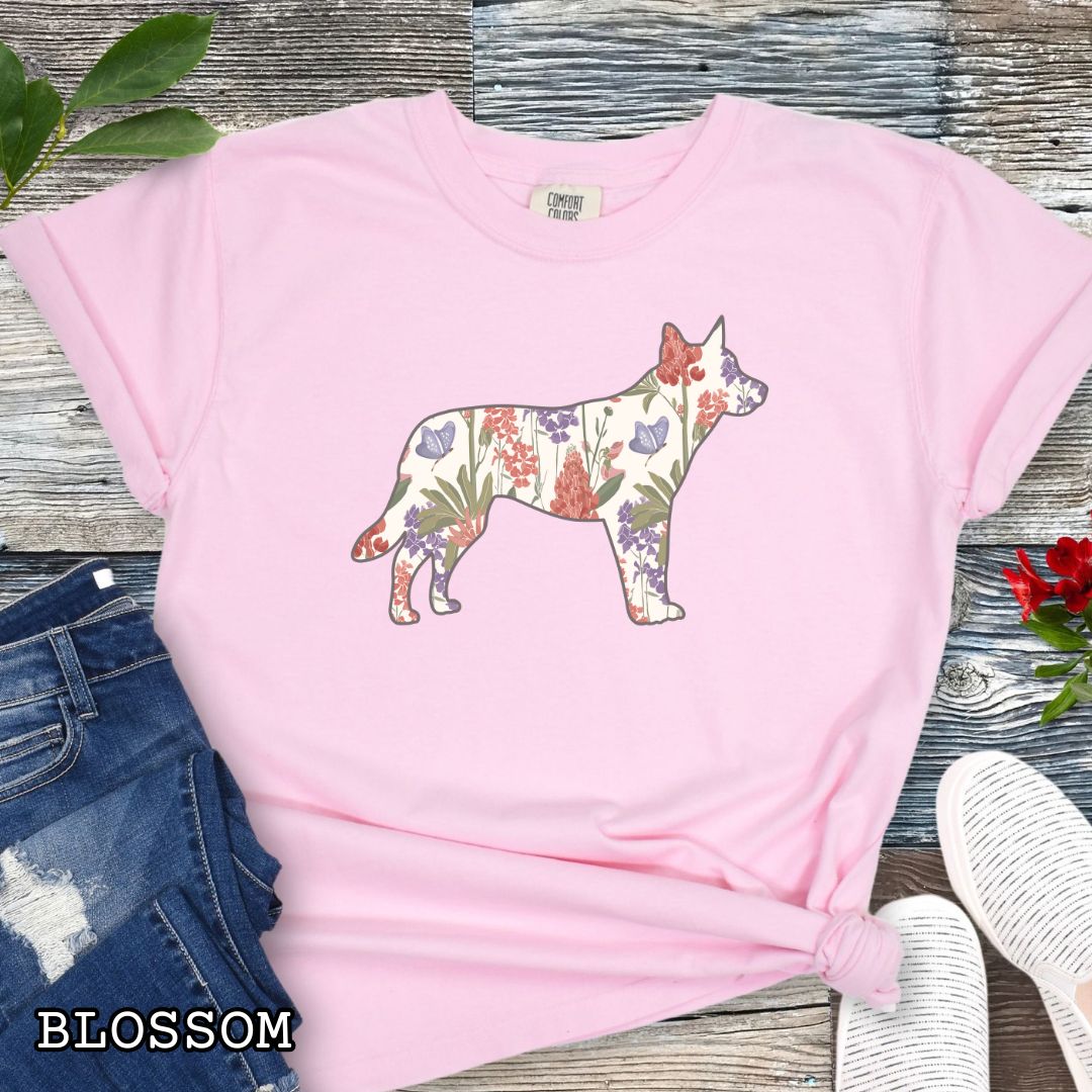 Pink Comfort Colors t-shirt with a floral-filled silhouette of a cattle dog, accompanied by a pair of rolled-up blue jeans and a pair of white shoes on a wooden surface.