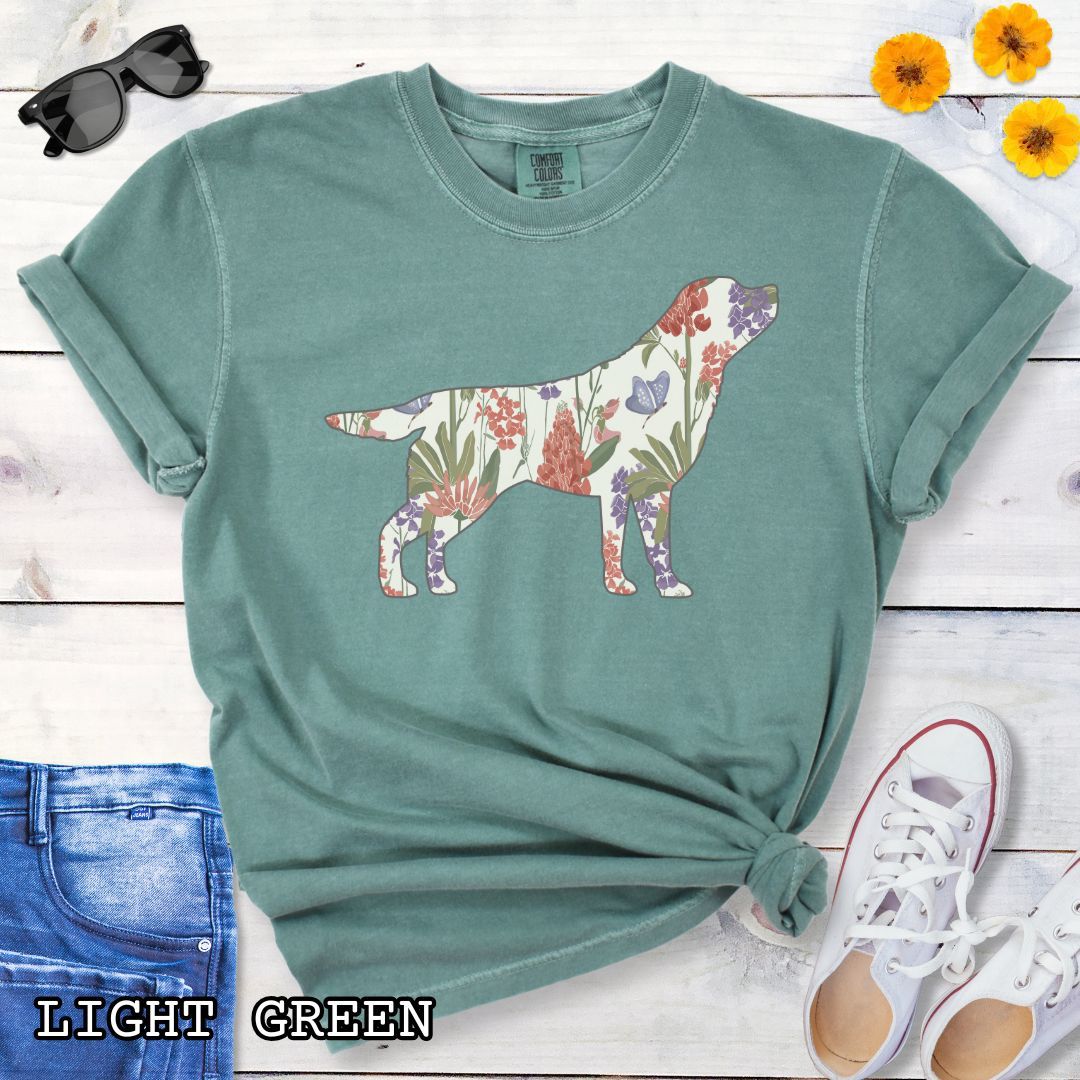 Light green Comfort Colors t-shirt with a floral-filled silhouette of a Labrador Retriever, accompanied by a pair of rolled-up blue jeans and a pair of white shoes on a wooden surface.