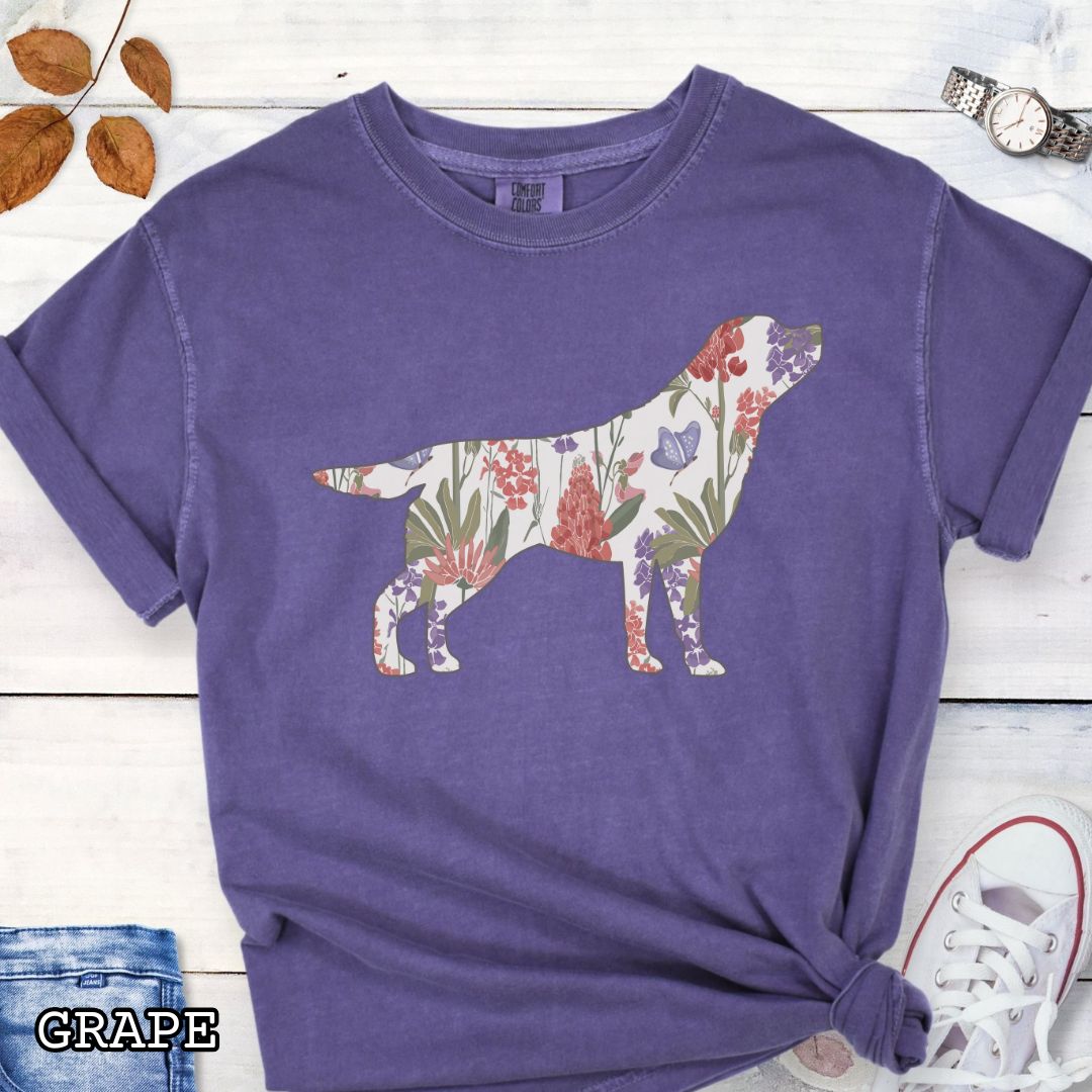 Purple Comfort Colors t-shirt with a floral-filled silhouette of a Labrador Retriever, accompanied by a pair of rolled-up blue jeans and a pair of white shoes on a wooden surface.