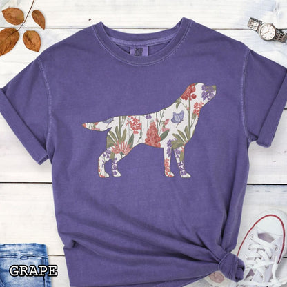 Purple Comfort Colors t-shirt with a floral-filled silhouette of a Labrador Retriever, accompanied by a pair of rolled-up blue jeans and a pair of white shoes on a wooden surface.