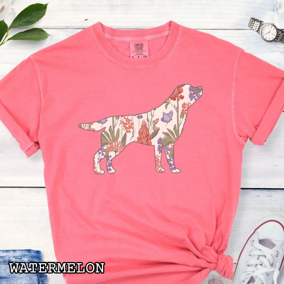 Watermelon-color Comfort Colors t-shirt with a floral-filled silhouette of a Labrador Retriever, accompanied by a pair of rolled-up blue jeans and a pair of white shoes on a wooden surface.