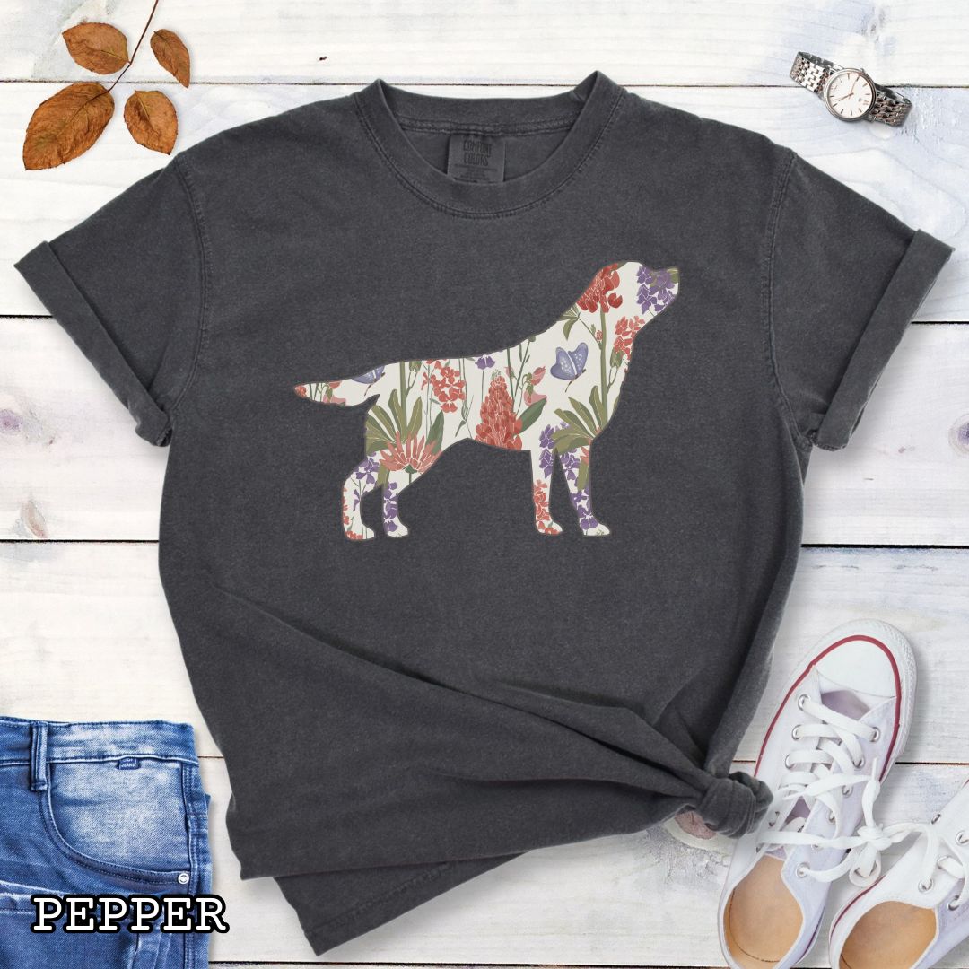 Dark grey Comfort Colors t-shirt with a floral-filled silhouette of a Labrador Retriever, accompanied by a pair of rolled-up blue jeans and a pair of white shoes on a wooden surface.