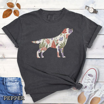 Dark grey Comfort Colors t-shirt with a floral-filled silhouette of a Labrador Retriever, accompanied by a pair of rolled-up blue jeans and a pair of white shoes on a wooden surface.