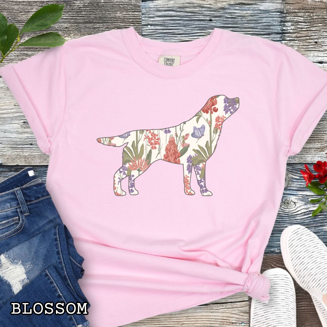 Pink Comfort Colors t-shirt with a floral-filled silhouette of a Labrador Retriever, accompanied by a pair of rolled-up blue jeans and a pair of white shoes on a wooden surface.