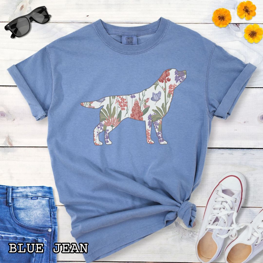 Blue Comfort Colors t-shirt with a floral-filled silhouette of a Labrador Retriever, accompanied by a pair of rolled-up blue jeans and a pair of white shoes on a wooden surface.