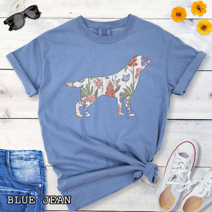 Blue Comfort Colors t-shirt with a floral-filled silhouette of a Labrador Retriever, accompanied by a pair of rolled-up blue jeans and a pair of white shoes on a wooden surface.
