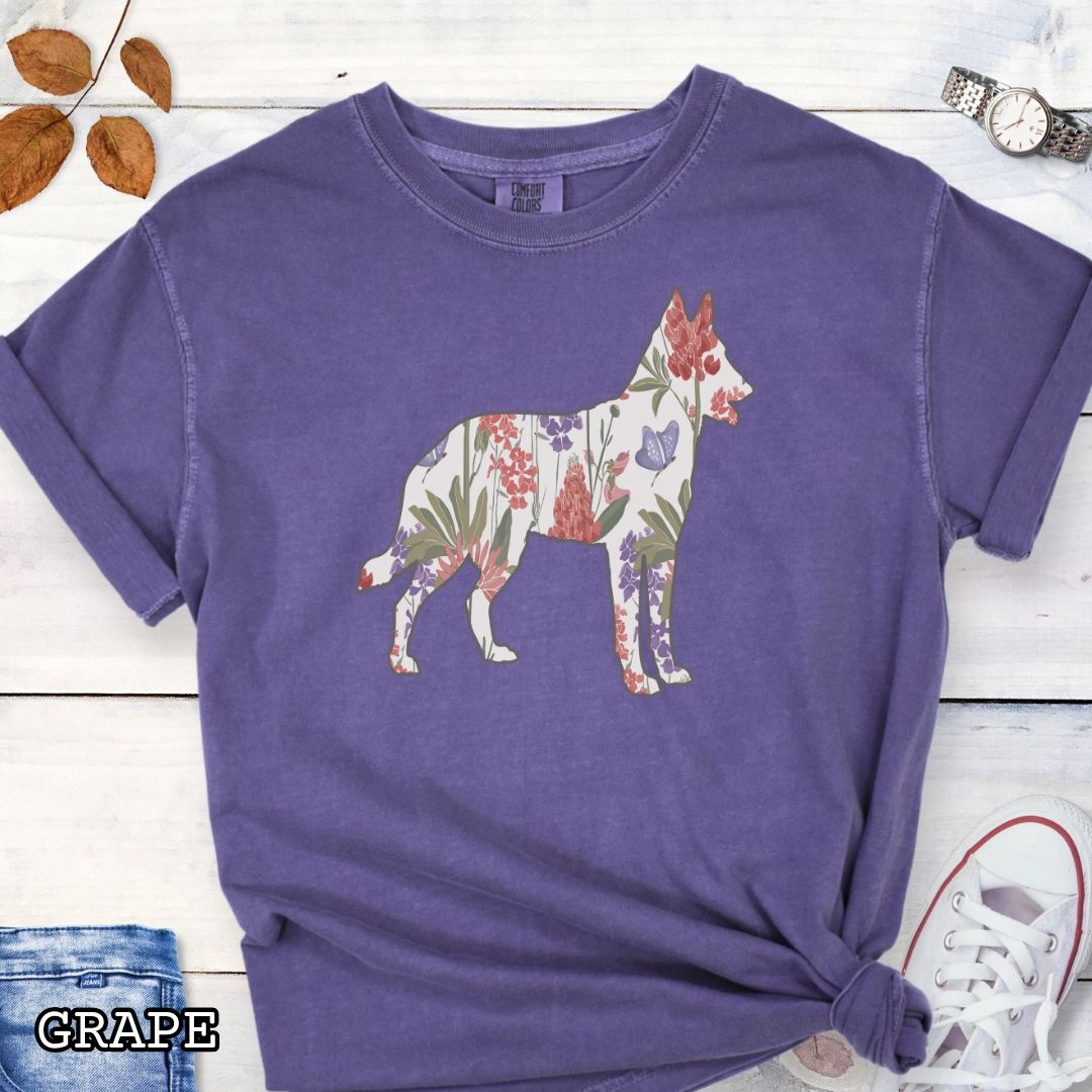 Purple Comfort Colors t-shirt with a floral-filled silhouette of a Belgian Malinois, accompanied by a pair of rolled-up blue jeans and a pair of white shoes on a wooden surface.