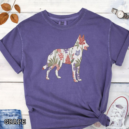Purple Comfort Colors t-shirt with a floral-filled silhouette of a Belgian Malinois, accompanied by a pair of rolled-up blue jeans and a pair of white shoes on a wooden surface.