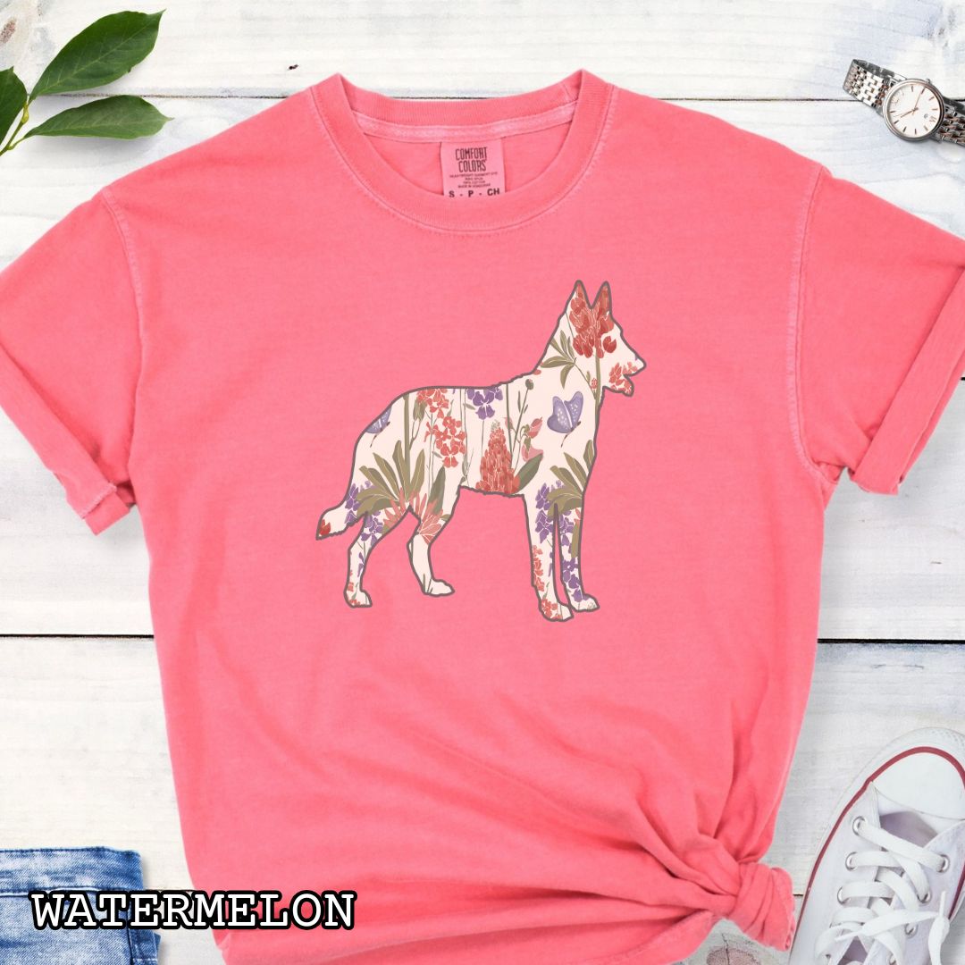 Watermelon-color Comfort Colors t-shirt with a floral-filled silhouette of a Belgian Malinois, accompanied by a pair of rolled-up blue jeans and a pair of white shoes on a wooden surface.