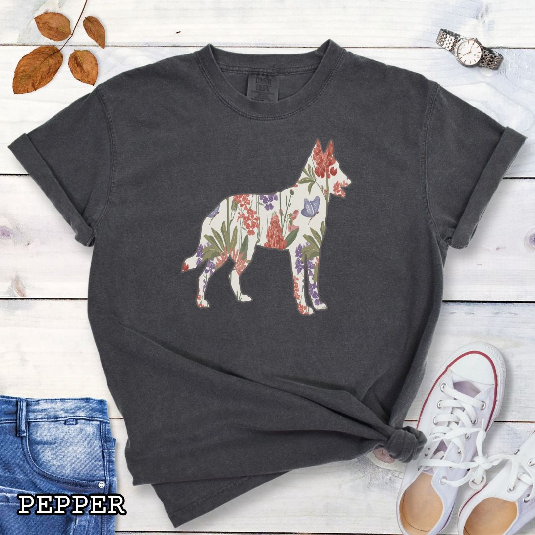 Dark grey Comfort Colors t-shirt with a floral-filled silhouette of a Belgian Malinois, accompanied by a pair of rolled-up blue jeans and a pair of white shoes on a wooden surface.