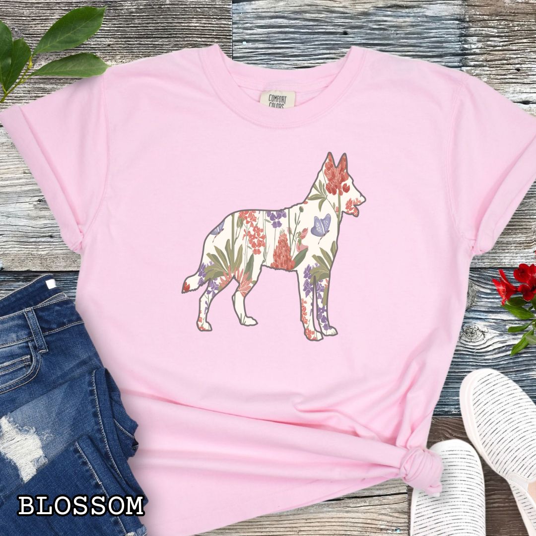 Pink Comfort Colors t-shirt with a floral-filled silhouette of a Belgian Malinois, accompanied by a pair of rolled-up blue jeans and a pair of white shoes on a wooden surface.