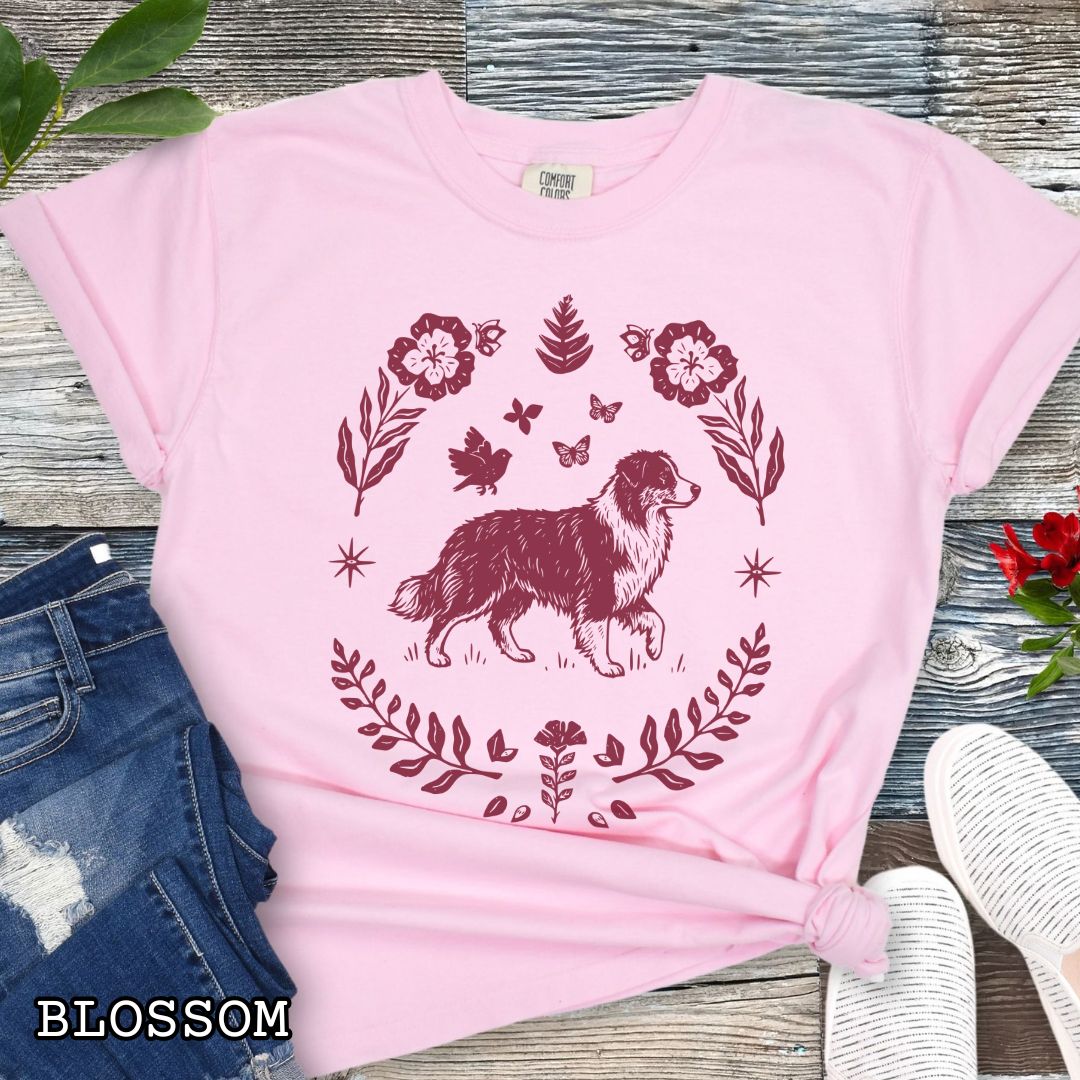 Pink Comfort Colors t-shirt for Australian Shepherd lovers with linocut-style monochrome design of an Aussie dog surrounded by floral botanical elements, shown on a wooden surface with jeans and flowers.