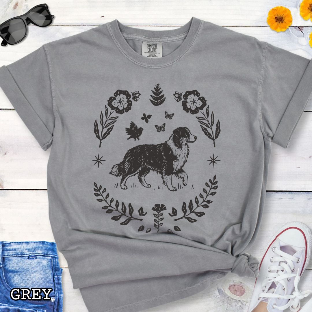 Grey Comfort Colors t-shirt for Australian Shepherd lovers with linocut-style monochrome design of an Aussie dog surrounded by floral botanical elements, shown on a wooden surface with jeans and flowers.