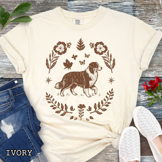 Beige Comfort Colors t-shirt for Australian Shepherd lovers with linocut-style monochrome design of an Aussie dog surrounded by floral botanical elements, shown on a wooden surface with jeans and flowers.