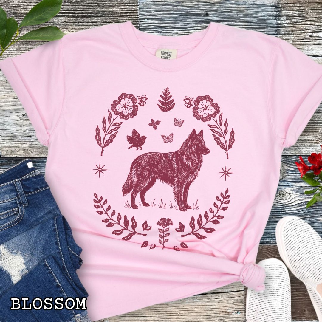 Pink Comfort Colors t-shirt for Belgian Sheepdog/Tervuren lovers, with folk art design of Belgian Shepherd surrounded by botanical elements, done in linocut monochrome style. Shown on a wooden surface accompanied by jeans, shoes and accessories.