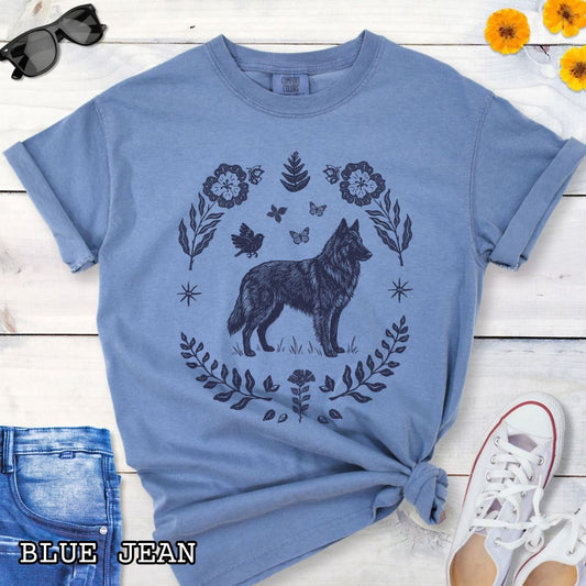 Blue Comfort Colors t-shirt for Belgian Sheepdog/Tervuren lovers, with folk art design of Belgian Shepherd surrounded by botanical elements, done in linocut monochrome style. Shown on a wooden surface accompanied by jeans, shoes and accessories.